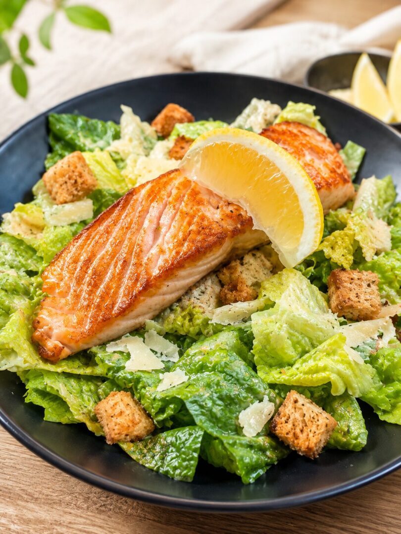 Plate of grilled salmon caesar salad with lemon wedges, placed on a wooden surface with a cloth